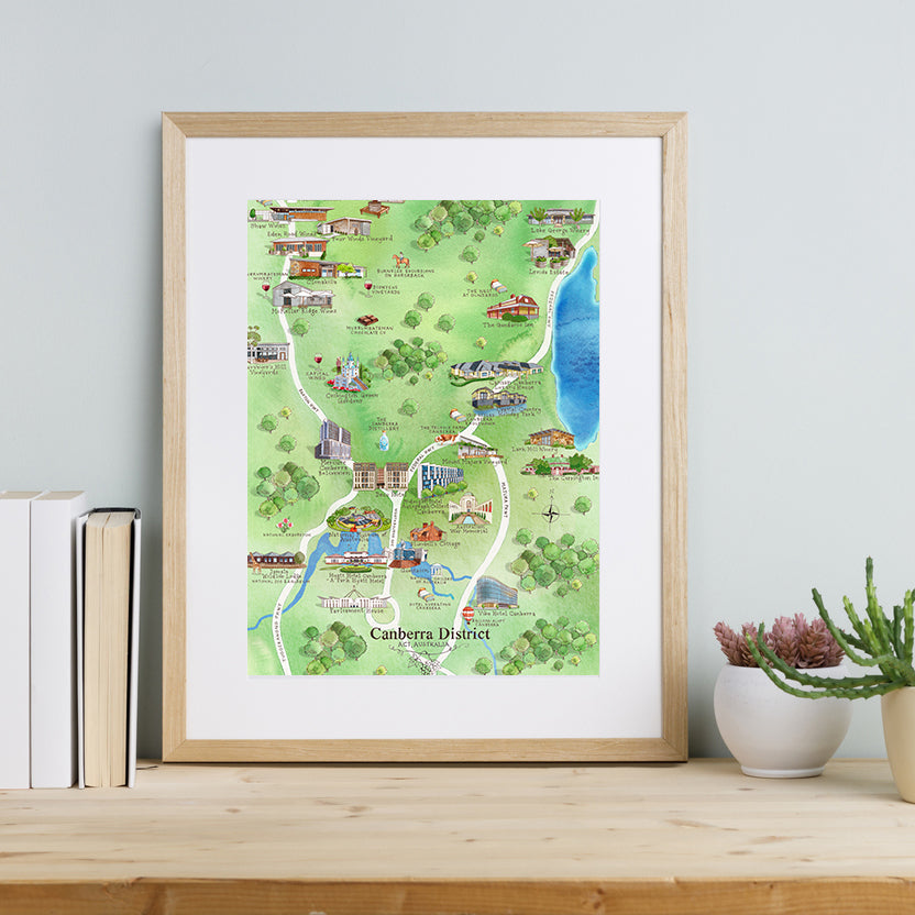 A framed A3-size art print of the Canberra District Wine Region Map, displayed on a wooden shelf with books and a plant to the side.