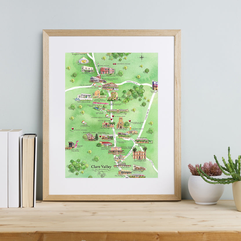A framed A3-size print of Clare Valley Wine Region Map, featuring illustrations of iconic wineries, accommodation, and local attractions, displayed on a wooden shelf with books and a plant to the side.