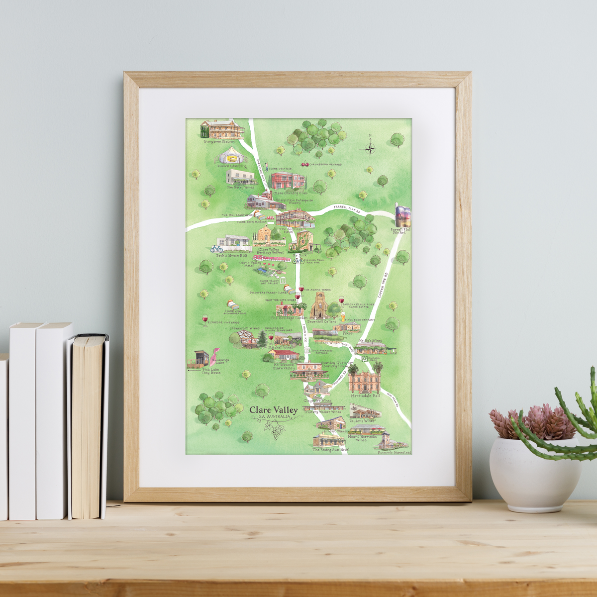 A framed A3-size print of Clare Valley Wine Region Map, featuring illustrations of iconic wineries, accommodation, and local attractions, displayed on a wooden shelf with books and a plant to the side
