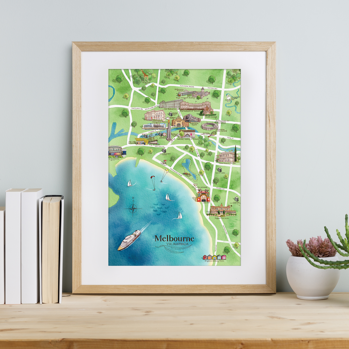 A framed A3-size print of Melbourne tourist attractions map, displayed on a wooden shelf with books and a plant to the side.