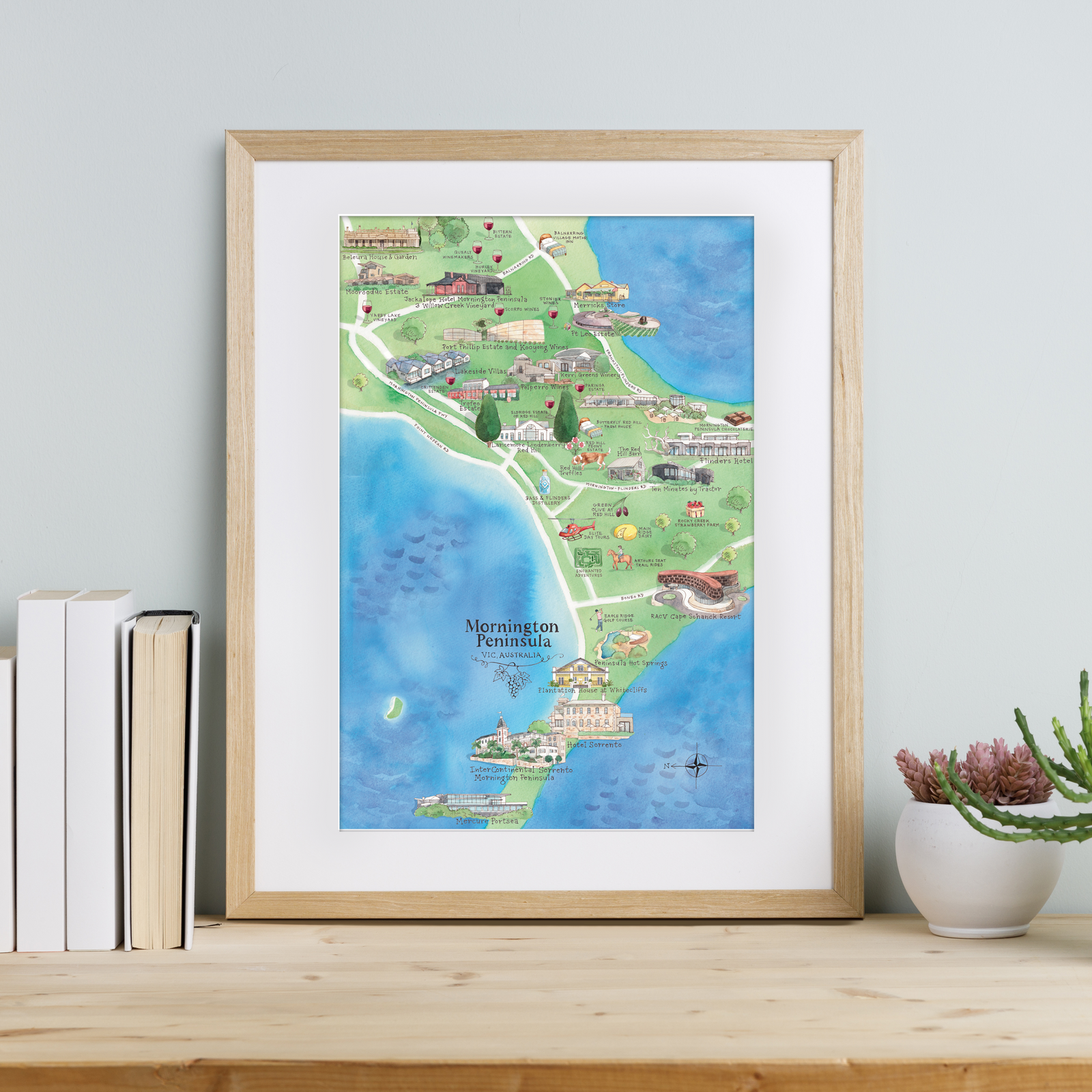 A framed A3-size print of the Mornington Peninsula wine region map, featuring iconic wineries, accommodation, and local attractions, displayed on a wooden shelf with books and a plant to the side.