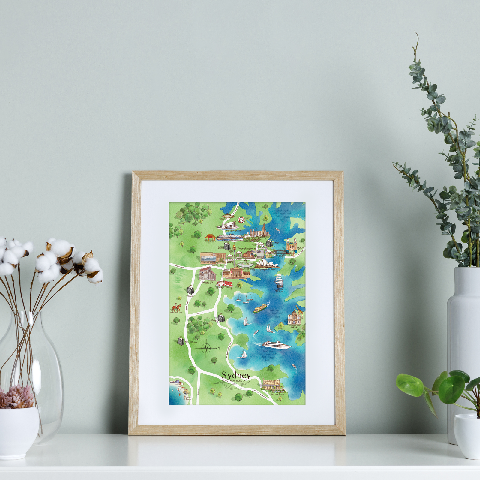 A framed A4-size print showcasing a colorful map of Sydney's tourist attractions, displayed on a shelf with decorative items.